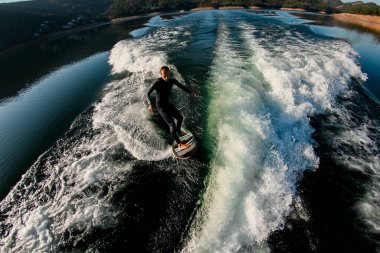 Siyah dalgıç kıyafetli genç bir sporcunun Wakesurf tahtasındaki dalgada ustaca dengede duruşu.