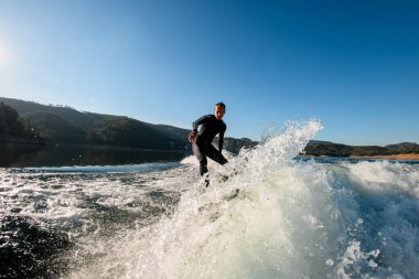 Siyah dalgıç kıyafetli genç adam uyanma tahtasındaki su sıçrama dalgası üzerinde enerjik bir şekilde dengede duruyor.