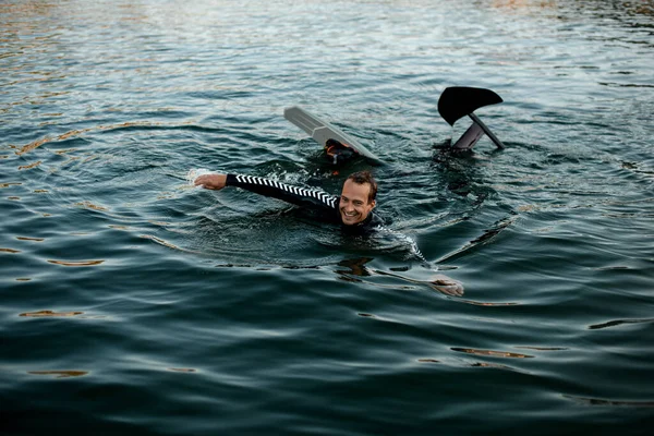 smiling man in a black wetsuit with a foil wakeboard swims in the water. Summertime watersports activity