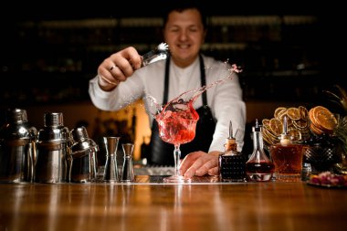 Bar tezgahında güzel bir manzara çeşitli bar ekipmanları ve şeffaf kristal kadehle barmenin içine barmenin buz küpünü fırlattığı bir içecek.