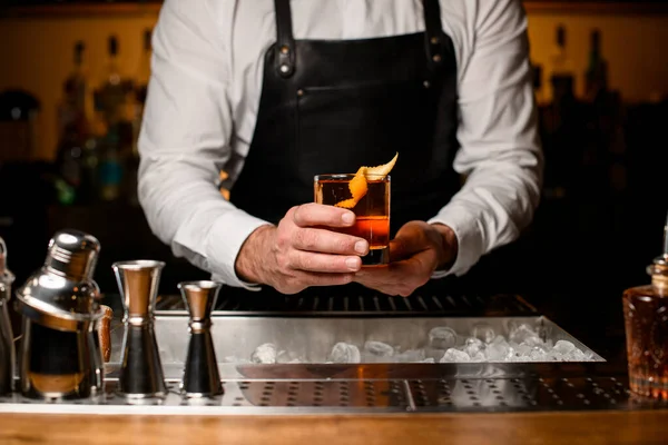 great view on hands of man bartender holding transparent glass with alcoholic drink with ice cube garnishing with orange zest