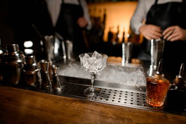 Bar tezgahında buz küpleri ve farklı bar ekipmanları olan kristal bir kadehe odaklanın.