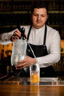 male bartender skilfully adds carbonated liquid from siphon to transparent glass with cold alcoholic cocktail on bar counter clipart