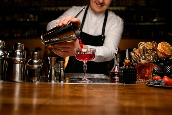 Bar tezgahında çeşitli bar ekipmanları ve şeffaf bir kristal kadehle barmen bir bardaktan kokteyl doldurur.
