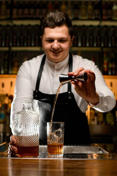 バーテンダーマンはジッガーを手に持ち氷のキューブで透明なガラスにドリンクを注ぐ バーでドリンクとクリスタルボトル — ストック写真