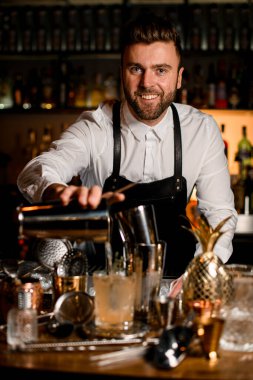 Çelik bardaktan kristal bardağa içki dolduran gülümseyen erkek barmenin yüzüne odaklan. Bar tezgahında farklı bar ekipmanları var.