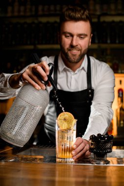 Erkek barmen sifondan şeffaf bardağa karbonatlı sıvı ekliyor ve tezgahta soğuk tazelik kokteyli var.