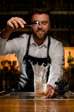 Barmenlerin maşadan bir parça buz attığı barın tezgahına su sıçratan şeffaf cam manzarası. Arkadaki barmen.