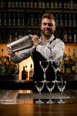 Yakışıklı gülümseyen barmen ellerinde büyük bir titretici ve tezgahta boş bardaklar piramidi tutuyor.