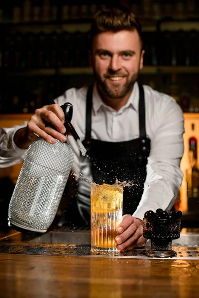 man bartender skilfully adds carbonated liquid from siphon to transparent glass with cold freshness cocktail on bar counter