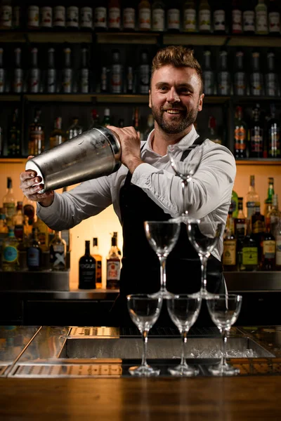 handsome smiling bartender holds large shaker in his hands and pyramid of empty glasses on bar counter on foreground