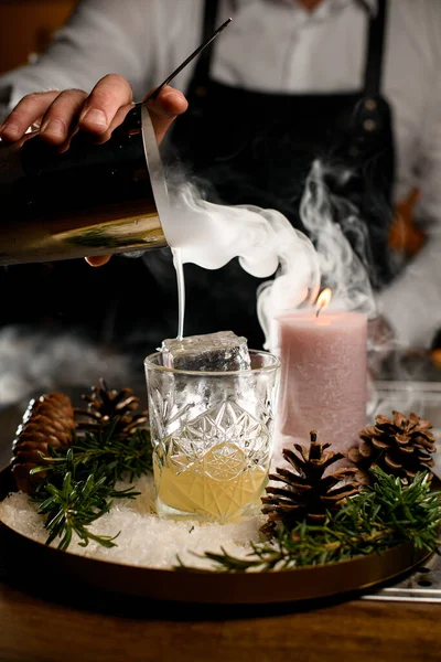 close-up of crystal glass with ice cubes in which pour a steaming drink from a steel shaker. Pine cones and a burning candle on tray near glass.
