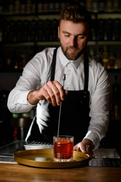 Man bartender masterfully mixing cold cocktail in a glass with a long bar spoon on bar