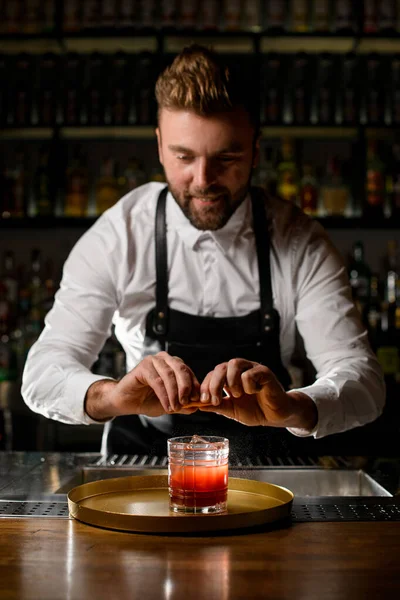 Man bartender gently sprinkles by citrus peel on a glass with a cocktail on a tray on the bar counter