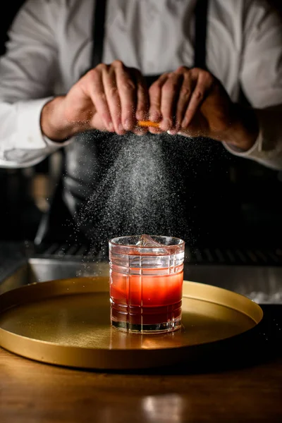 Close-up of glass with alcoholic beverage stand on bar while hands of male bartender carefully sprinkle by citrus zest juice on it