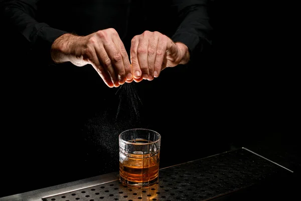 Close-up view of hands of man bartender spraying oils from an orange peel on cocktail on dark background.