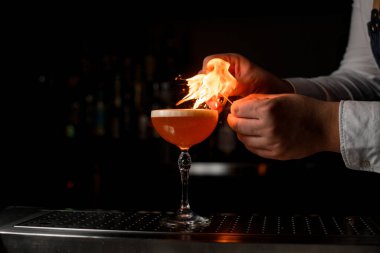 Çelik tezgahın üzerinde ekşi alkollü kokteyl bulunan şarap kadehinin yakın çekimi ve barmenin elleri üzerine ateş yakar.