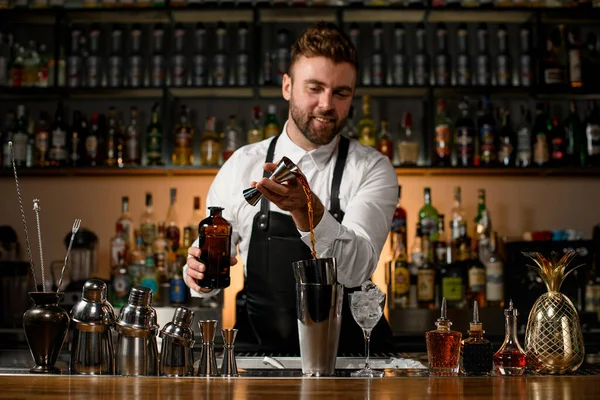 various steel shakers and bottles stand on the bar counter, and man bartender gently pours an alcoholic drink from jigger in the shaker cup