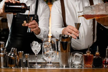 İki erkek barmenin, ellerinde şişeler ve içecekleri çelik zımbırtılara dökerken yakın plan görüntüsü. Çalkalayıcılar ve şişeler ön plandaki tezgahın üzerinde sıraya dizilir.