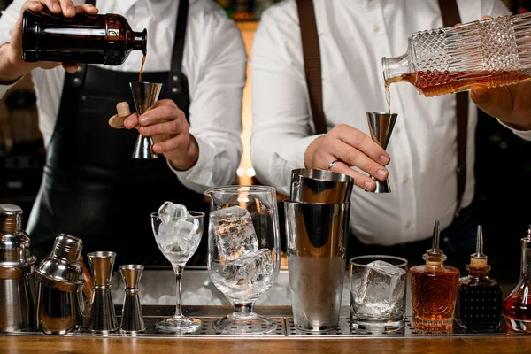 Close-up view of two male bartenders holding bottles and pours beverage into steel jiggers. Different shakers and bottles stand in a row on the bar counter in the foreground