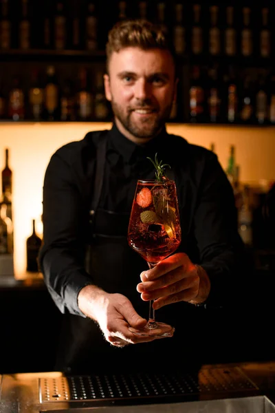 handsome man bartender gently holds glass with cold alcoholic drink with strawberries and mint decorated with branch of rosemary