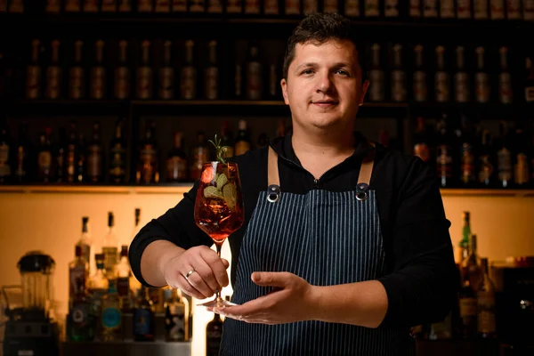 Man bartender gently holds glass with cold alcoholic drink with strawberries and mint decorated with branch of rosemary