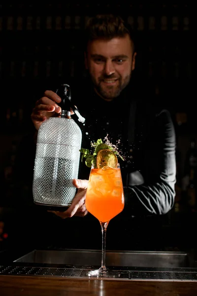 beautiful glass with bright orange drink and ice garnished with fresh mint leaves on which male bartender sprinkles from a siphon