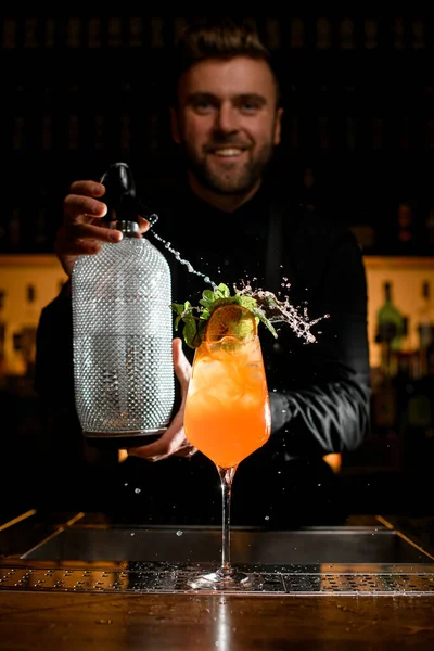 glass with bright orange drink and ice garnished with fresh mint leaves on which male bartender sprinkles from a siphon