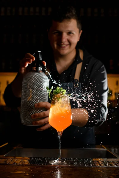 wineglass with bright orange drink and ice garnished with fresh mint leaves on which the bartender sprinkles from a siphon