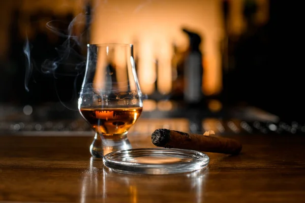 Selective focus on smoking cigar on ashtray and glass with whiskey on blurred bar background