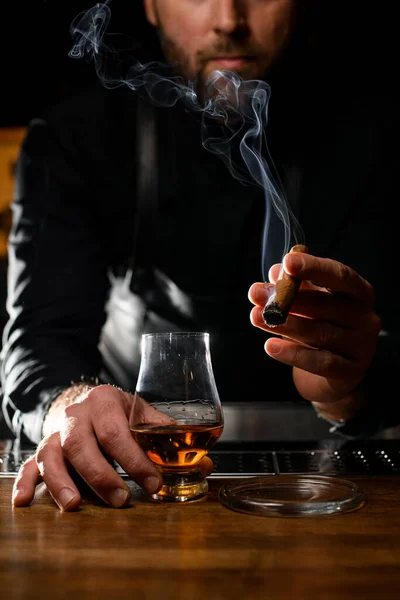 smoking cuban cigar in the hand of a male bartender and a glass of cognac on the bar counter