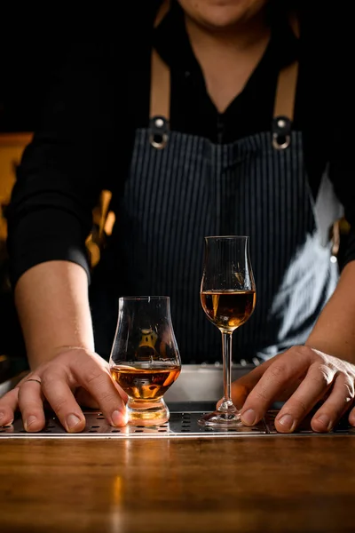 two beautiful glasses with a strong alcoholic drink stand on the bar and the hands of male bartender nearby