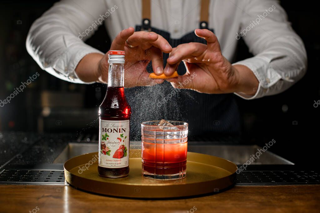 UKRAINE, KYIV - DECEMBER 06, 2021: close-up of a bottle of monin syrup and a glass of drink on tray and male hands splashing by orange zest