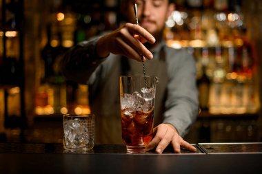 hand of bartender hold long bar spoon and stirring drink in glass with ice. Crystal glass with ice cubes nearby on bar counter. Blurry bartender in the background
