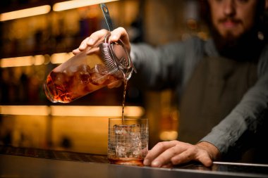 Hand of bartender holds transparent mixing cup with ice cubes and drink with strainer and poured drink into glass on bar