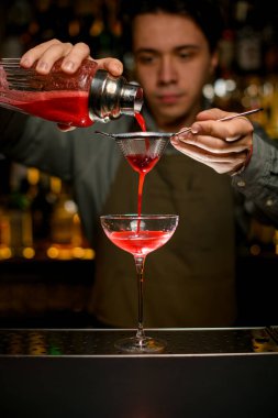 hand of male bartender holds sieve and pours cocktail through it from glass shaker into wine glass on bar counter