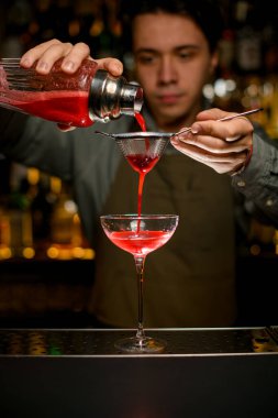 hand of male bartender holds sieve and pours cocktail through it from glass shaker into wine glass on bar counter clipart
