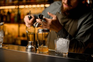 Close-up view on glassy shaker into which hand of the male bartender pouring drink from steel jigger