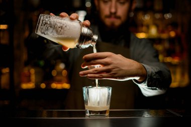 hands of male bartender gently holds shaker and pours cold cocktail into glass filtering it through sieve