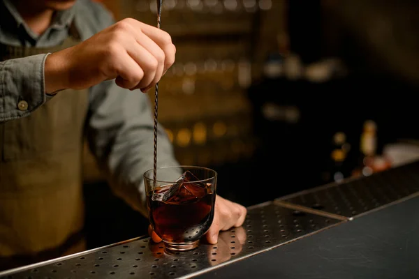 hand of bartender hold long bar spoon and gently stirring cold drink in glass on bar. Blurred background