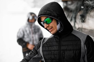 portrait of young man in sunglasses in black jacket with hood on his head on blurred background