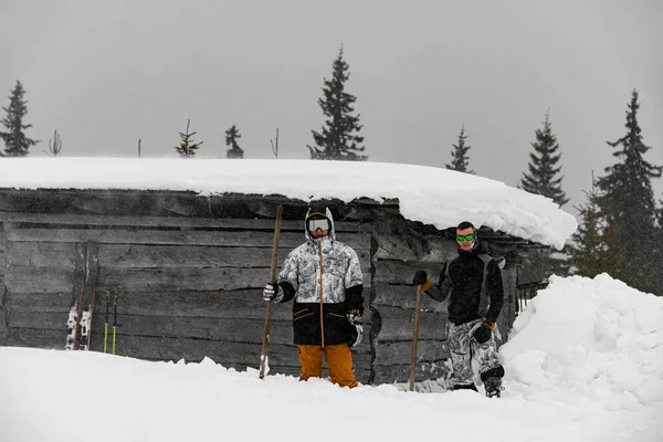 view on two men in ski suit and ski mask with shovels in hands on background of snow covered wooden ski hut
