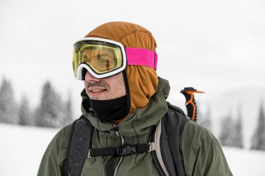 Portrait of male skier in ski mask on his head against the backdrop of blurred evergreen trees and white snow