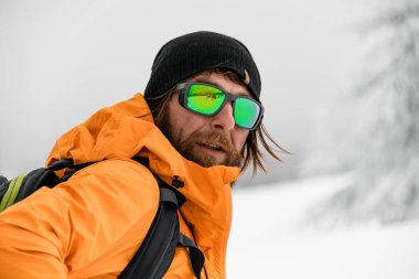 Close-up portrait of bearded man in bright jacket and black hat with glasses with reflection of winter ski resort.
