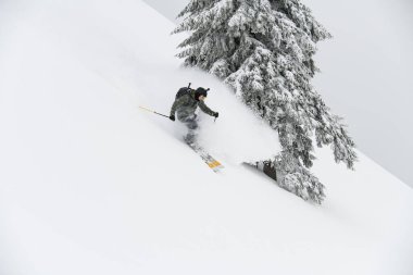 Male freerider skier with go-pro camera on his helmet fast sliding down snow covered slope. Beautiful fir tree on background
