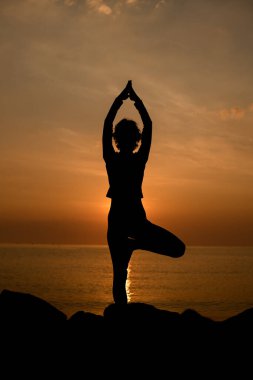 Dark silhouette of woman who practices yoga on the beach, standing in a tree pose at sunset. Healthy lifestyle concept.