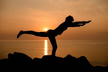Balancing Stick Pose. Tuladandasana. Side view of woman silhouette doing yoga or gymnastics outdoor at morning. Morning sky with sun reflecting in water in background