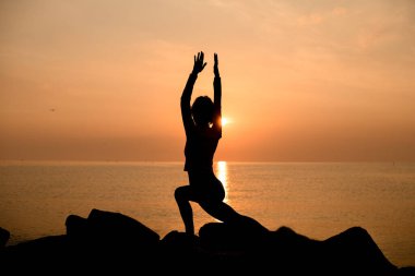 Silhouette of woman practicing yoga or pilates at sunset or sunrise doing lunge exercise standing in Warrior pose. Virabhadrasana