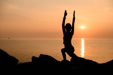 Dark silhouette of woman practicing yoga or pilates at sunset or sunrise doing lunge exercise standing in Warrior pose. Virabhadrasana