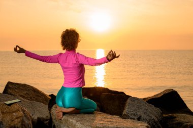 rear view of woman sitting on her knees with her arms outstretched against the backdrop of the sea and sunset. Yoga pose.